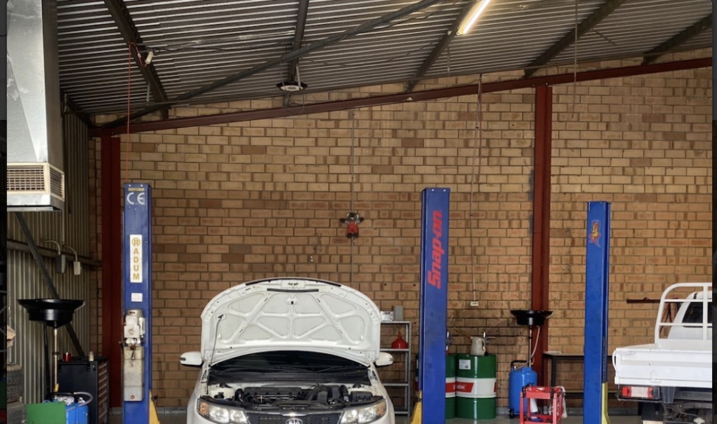 A white sedan up on a two-post hoist in the brick-walled workshop, bonnet open